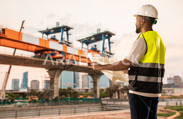 Concept of architectural engineering and construction, study of the basics of the construction plan, work in the engineering sector, a Saudi Arabian Gulf engineer wearing a protective vest and helmet holding construction plans in his hands, reviewing and presenting project manuscripts in full, project management and follow-up