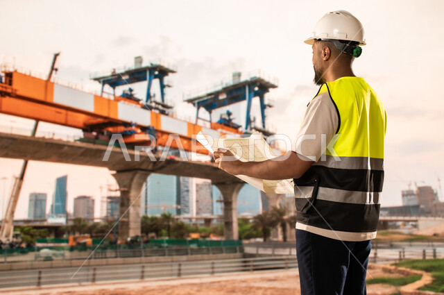 Concept of architectural engineering and construction, study of the basics of the construction plan, work in the engineering sector, a Saudi Arabian Gulf engineer wearing a protective vest and helmet holding construction plans in his hands, reviewing and presenting project manuscripts in full, project management and follow-up