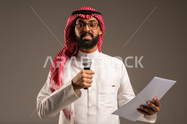 Reading notes and information, having dialogues and discussions, close-up portrait of a Saudi Arabian Gulf man wearing traditional thobe and shemagh holding a bunch of papers and a loudspeaker giving speeches and looking at the camera, preparing and arranging speech ideas in advance, gray background