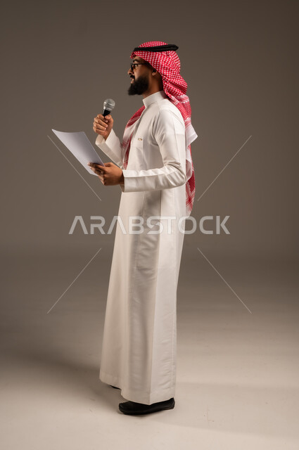 إعداد وتحضير أفكار الخطاب بشكل مسبق، القيام بالحوارات والنقاشات، بورتريه من الجانب لرجل عربي خليجي سعودي يرتدي الثوب التقليدي والشماغ يحمل بيده مجموعة من الأوراق ومكبر الصوت يقوم بإلقاء الخطابات، صورة الجسم بالطول الكامل، خلفية رمادية