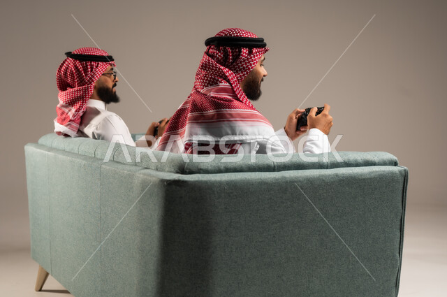 المشاركة والمنافسة بالألعاب التقنية الحديثة والمتطورة,  بورتريه لرجلين عربيين خليجيين سعوديين يرتديان الثوب التقليدي والشماغ يمسكان عصا التحكم, العالم الرقمي الترفيهي, المتعة والتسلية بالفيديو جيمز برفقة الأصدقاء
