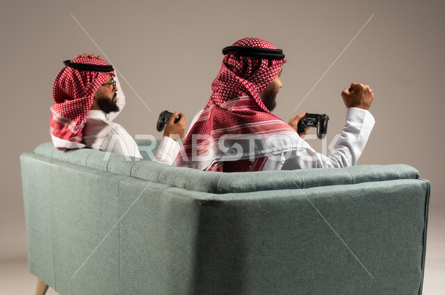 المشاركة والمنافسة بالألعاب التقنية الحديثة والمتطورة,  بورتريه لرجلين عربيين خليجيين سعوديين يرتديان الثوب التقليدي والشماغ يمسكان عصا التحكم, العالم الرقمي الترفيهي, المتعة والتسلية بالفيديو جيمز برفقة الأصدقاء