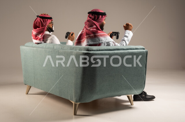 المشاركة والمنافسة بالألعاب التقنية الحديثة والمتطورة,  بورتريه لرجلين عربيين خليجيين سعوديين يرتديان الثوب التقليدي والشماغ يمسكان عصا التحكم, العالم الرقمي الترفيهي, المتعة والتسلية بالفيديو جيمز برفقة الأصدقاء