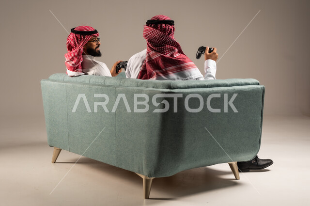 المشاركة والمنافسة بالألعاب التقنية الحديثة والمتطورة,  بورتريه لرجلين عربيين خليجيين سعوديين يرتديان الثوب التقليدي والشماغ يمسكان عصا التحكم, العالم الرقمي الترفيهي, المتعة والتسلية بالفيديو جيمز برفقة الأصدقاء