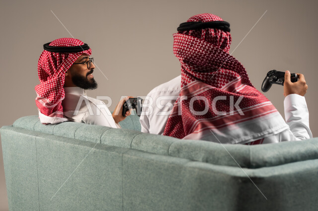 المشاركة والمنافسة بالألعاب التقنية الحديثة والمتطورة,  بورتريه لرجلين عربيين خليجيين سعوديين يرتديان الثوب التقليدي والشماغ يمسكان عصا التحكم, العالم الرقمي الترفيهي, المتعة والتسلية بالفيديو جيمز برفقة الأصدقاء