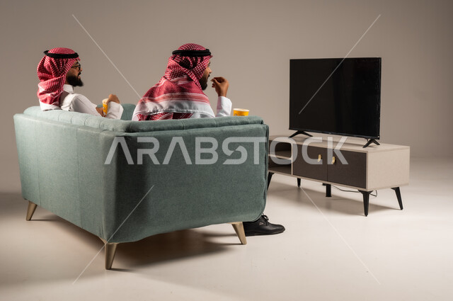 مفهوم التسليه والترفيه الجماعي, قضاء وقت ممتع بتناول الفوشار اثناء مشاهده التلفاز، بورتريه لصديقين عربيين خليجيين سعوديين يرتديان الشماغ والثوب التقليدي يجلسان على الأريكه يحملان بيدهما أكواب مليئه بالبوب كورن، تعابير وايماءات السعاده والسرور، خلفية رمادية