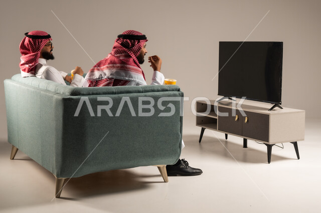 مفهوم التسليه والترفيه الجماعي, قضاء وقت ممتع بتناول الفوشار اثناء مشاهده التلفاز، بورتريه لصديقين عربيين خليجيين سعوديين يرتديان الشماغ والثوب التقليدي يجلسان على الأريكه يحملان بيدهما أكواب مليئه بالبوب كورن، تعابير وايماءات السعاده والسرور، خلفية رمادية