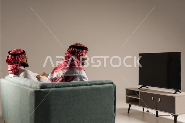 مفهوم التسليه والترفيه الجماعي, قضاء وقت ممتع بتناول الفوشار اثناء مشاهده التلفاز، بورتريه لصديقين عربيين خليجيين سعوديين يرتديان الشماغ والثوب التقليدي يجلسان على الأريكه يحملان بيدهما أكواب مليئه بالبوب كورن، تعابير وايماءات السعاده والسرور، خلفية رمادية