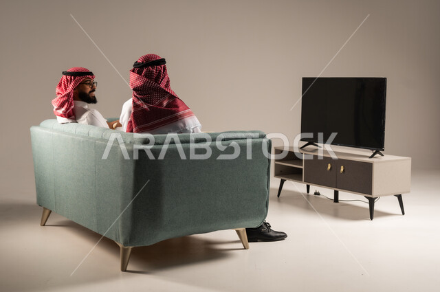 مفهوم التسليه والترفيه الجماعي, قضاء وقت ممتع بتناول الفوشار اثناء مشاهده التلفاز، بورتريه لصديقين عربيين خليجيين سعوديين يرتديان الشماغ والثوب التقليدي يجلسان على الأريكه يحملان بيدهما أكواب مليئه بالبوب كورن، تعابير وايماءات السعاده والسرور، خلفية رمادية