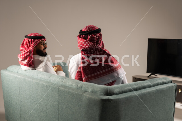 مفهوم التسليه والترفيه الجماعي, قضاء وقت ممتع بتناول الفوشار اثناء مشاهده التلفاز، بورتريه لصديقين عربيين خليجيين سعوديين يرتديان الشماغ والثوب التقليدي يجلسان على الأريكه يحملان بيدهما أكواب مليئه بالبوب كورن، تعابير وايماءات السعاده والسرور، خلفية رمادية