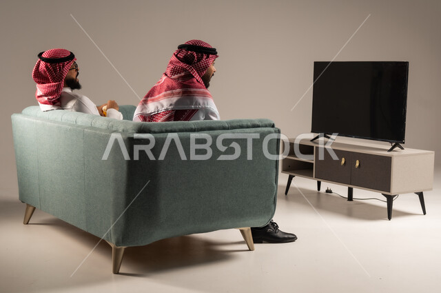 قضاء وقت ممتع بتناول الفوشار اثناء مشاهده التلفاز، مفهوم التسليه والترفيه الجماعي, بورتريه لصديقين عربيين خليجيين سعوديين يرتديان الشماغ والثوب التقليدي يجلسان على الأريكه يحملان بيدهما أكواب مليئه بالبوب كورن، ايماءات السعاده والسرور، خلفية رمادية