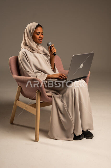 استخدام التقنيات التكنولوجية الحديثة، القراءة من الحاسوب المحمول، تقديم العروض الفنيه والغنائيه والفقرات الترفيهية، بورتريه لشابة عربية خليجية سعودية محجبة ترتدي العباءة الملونة تمسك بيدها المايكروفون بإيماءات الاندماج بالغناء، خلفيه رمادية