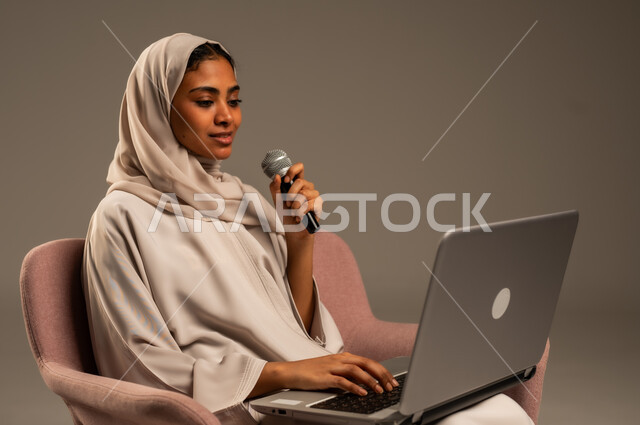 استخدام التقنيات التكنولوجية الحديثة، القراءة من الحاسوب المحمول، تقديم العروض الفنيه والغنائيه والفقرات الترفيهية، بورتريه لشابة عربية خليجية سعودية محجبة ترتدي العباءة الملونة تمسك بيدها المايكروفون بإيماءات الاندماج بالغناء، خلفيه رمادية