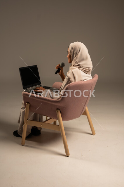 Using modern technological techniques, reading from a laptop, presenting artistic and singing performances and entertainment segments, a portrait of a veiled Saudi Arabian Gulf young woman wearing a colorful abaya holding a microphone in her hand with gestures of integration into singing, gray background