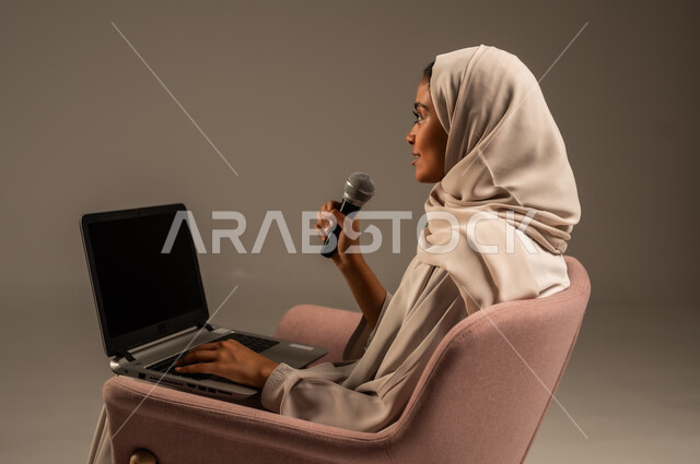 Using modern technological techniques, reading from a laptop, presenting artistic and singing performances and entertainment segments, a portrait of a veiled Saudi Arabian Gulf young woman wearing a colorful abaya holding a microphone in her hand with gestures of integration into singing, gray background