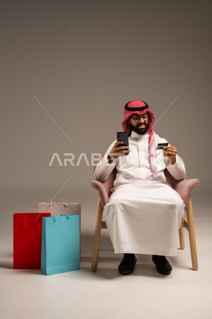 الشراء والدفع عن طريق بطاقة الإئتمان البنكية،  إتمام عملية الطلبات باستخدام الهاتف المحمول، مفهوم التسوق عن بعد, بورتريه لشاب عربي خليجي سعودي يرتدي الثوب التقليدي والشماغ يمسك بيده أكياس ورقية ملونة, التبضع والاستفادة من العروض والتخفيضات, خلفية رمادية