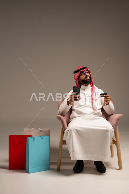 الشراء والدفع عن طريق بطاقة الإئتمان البنكية،  إتمام عملية الطلبات باستخدام الهاتف المحمول، مفهوم التسوق عن بعد, بورتريه لشاب عربي خليجي سعودي يرتدي الثوب التقليدي والشماغ يمسك بيده أكياس ورقية ملونة, التبضع والاستفادة من العروض والتخفيضات, خلفية رمادية