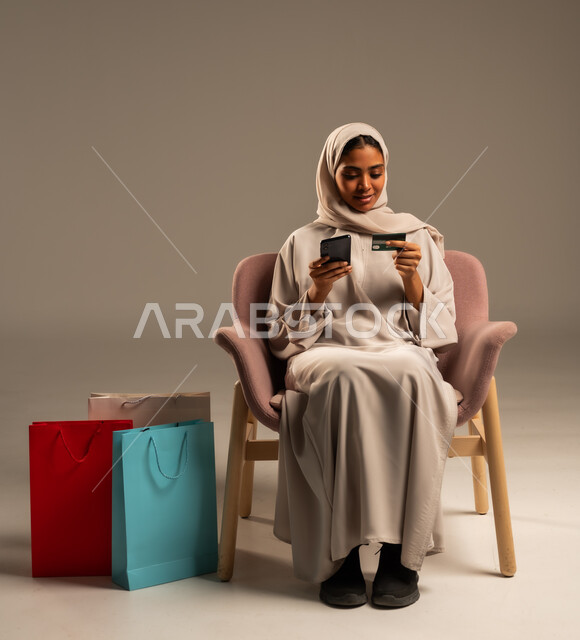 الدفع والشراء عن طريق بطاقة الإئتمان البنكية،  إتمام عملية الطلبات باستخدام الهاتف المحمول، مفهوم التسوق عن بعد, بورتريه  لإمرأة عربية خليجية سعودية ترتدي الحجاب والعباءة تحمل بيدها أكياس كرتون ملونه، التبضع والاستفادة من العروض والتخفيضات, خلفية رمادية