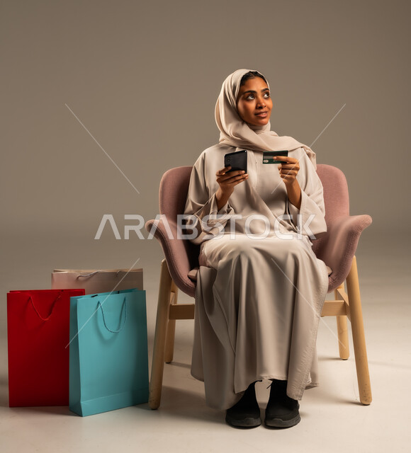 الدفع والشراء عن طريق بطاقة الإئتمان البنكية،  إتمام عملية الطلبات باستخدام الهاتف المحمول، مفهوم التسوق عن بعد, بورتريه  لإمرأة عربية خليجية سعودية ترتدي الحجاب والعباءة تحمل بيدها أكياس كرتون ملونه، التبضع والاستفادة من العروض والتخفيضات, خلفية رمادية