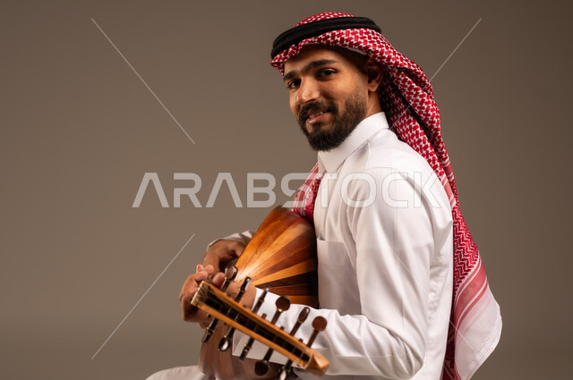 ممارسة الهوايات الفنية، فنان يقدم البرامج والفنون الموسيقية والفن المسرحي، مواهب فنية موسيقية، بورتريه لرجل خليجي سعودي عربي يرتدي الثوب التقليدي والشماغ يعزف على الة العود، الإستمتاع بالعزف على الآلات الوترية الكلاسيكية، جلسة وناسة وطرب، خلفية رمادية
