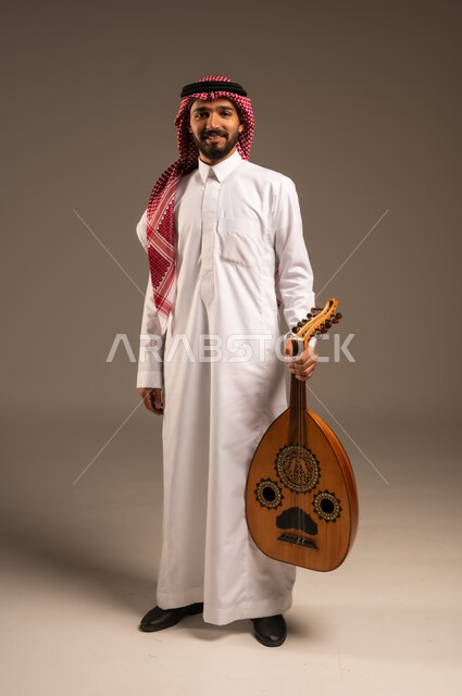 ممارسة الهوايات الفنية، فنان يقدم البرامج والفنون الموسيقية والفن المسرحي، مواهب فنية موسيقية، بورتريه لرجل خليجي سعودي عربي يرتدي الثوب التقليدي والشماغ يعزف على الة العود، الإستمتاع بالعزف على الآلات الوترية الكلاسيكية، جلسة وناسة وطرب، خلفية رمادية