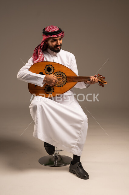 الإستمتاع بالعزف على الآلات الوترية الكلاسيكية، جلسة وناسة وطرب، ممارسة الهوايات الفنية، فنان يقدم البرامج والفنون الموسيقية والفن المسرحي، مواهب فنية موسيقية، بورتريه لرجل خليجي سعودي عربي يرتدي الثوب التقليدي والشماغ يعزف على الة العود، خلفية رمادية