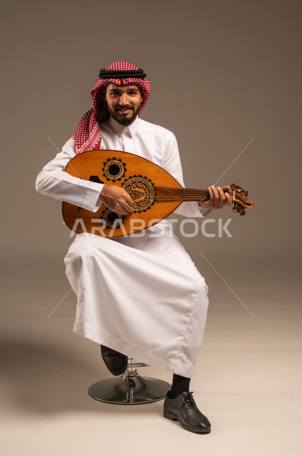 جلسة وناسة وطرب، ممارسة الهوايات الفنية، فنان يقدم البرامج والفنون الموسيقية والفن المسرحي، مواهب فنية موسيقية، بورتريه لرجل خليجي سعودي عربي يرتدي الثوب التقليدي والشماغ يعزف على الة العود، الإستمتاع بالعزف على الآلات الوترية الكلاسيكية، خلفية رمادية