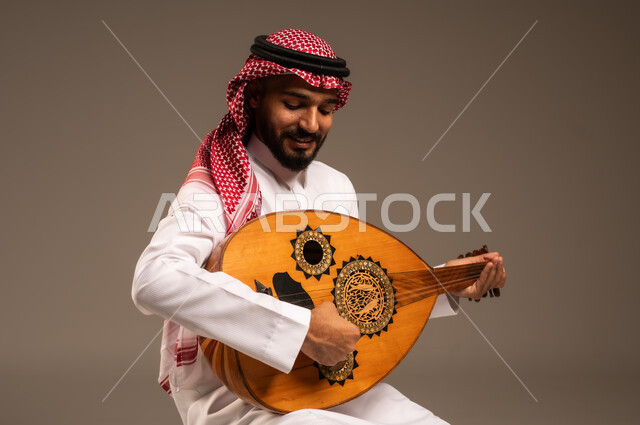 جلسة وناسة وطرب، ممارسة الهوايات الفنية، فنان يقدم البرامج والفنون الموسيقية والفن المسرحي، مواهب فنية موسيقية، بورتريه لرجل خليجي سعودي عربي يرتدي الثوب التقليدي والشماغ يعزف على الة العود، الإستمتاع بالعزف على الآلات الوترية الكلاسيكية، خلفية رمادية