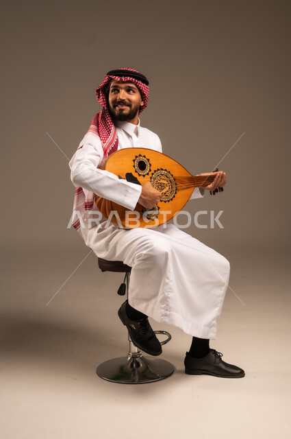 جلسة وناسة وطرب، ممارسة الهوايات الفنية، فنان يقدم البرامج والفنون الموسيقية والفن المسرحي، مواهب فنية موسيقية، بورتريه لرجل خليجي سعودي عربي يرتدي الثوب التقليدي والشماغ يعزف على الة العود، الإستمتاع بالعزف على الآلات الوترية الكلاسيكية، خلفية رمادية