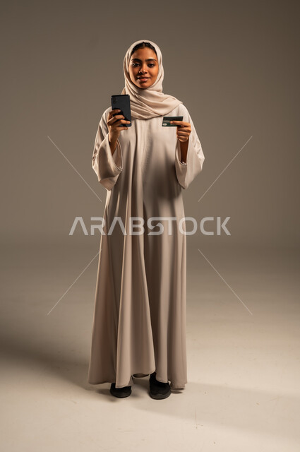 إمكانية الشراء والدفع من خلال برامج وتطبيقات عبر الجوال، بورتريه لسيدة عربية خليجية سعودية محجبة ترتدي عباءة ملونة تحمل بيدها الهاتف المحمول وبطاقة الإئتمان البنكية للتسوق الالكتروني، استخدام الاجهزة التقنية الحديثة، الدفع عن بعد عبر الانترنت، خلفية رمادية