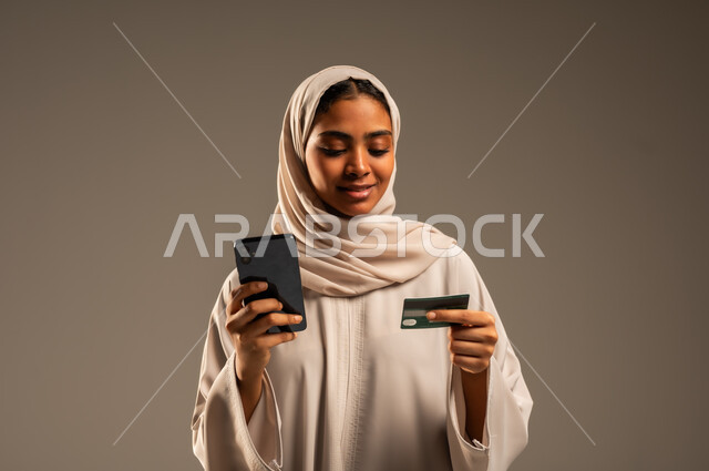 Possibility of purchasing and paying through mobile programs and applications, portrait of a veiled Saudi Arabian Gulf woman wearing a colorful abaya holding a mobile phone and a bank credit card for online shopping, using modern technical devices, remote payment via the Internet, gray background