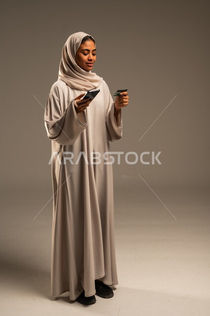 Possibility of purchasing and paying through mobile programs and applications, portrait of a veiled Saudi Arabian Gulf woman wearing a colorful abaya holding a mobile phone and a bank credit card for online shopping, using modern technical devices, remote payment via the Internet, gray background