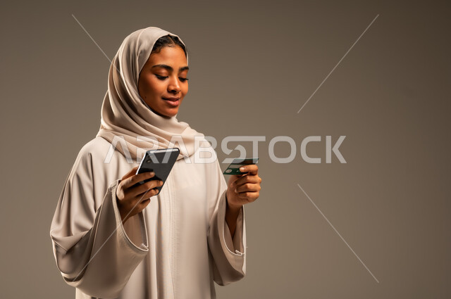 إمكانية الشراء والدفع من خلال برامج وتطبيقات عبر الجوال، بورتريه لسيدة عربية خليجية سعودية محجبة ترتدي عباءة ملونة تحمل بيدها الهاتف المحمول وبطاقة الإئتمان البنكية للتسوق الالكتروني، استخدام الاجهزة التقنية الحديثة، الدفع عن بعد عبر الانترنت، خلفية رمادية