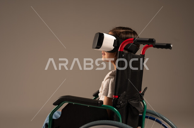  تحسين جودة حياة ذوي الاعاقة الحركية، استخدام كراسي المعاقين للمساعدة على الحركة والتنقل، بورتريه لفتاة عربية  خليجية سعودية ترتدي الزي الكاجوال تقوم بمشاهدة أفلام عبر نظارات الواقع الافتراضي ثلاثية الأبعاد، محاكاة العالم الخيالي والتفاعل مع الفيلم، خلفية رمادية