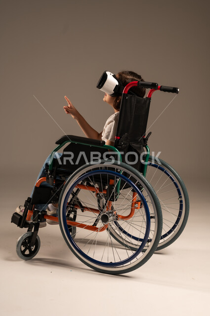  تحسين جودة حياة ذوي الاعاقة الحركية، استخدام كراسي المعاقين للمساعدة على الحركة والتنقل، بورتريه لفتاة عربية  خليجية سعودية ترتدي الزي الكاجوال تقوم بمشاهدة أفلام عبر نظارات الواقع الافتراضي ثلاثية الأبعاد، محاكاة العالم الخيالي والتفاعل مع الفيلم، خلفية رمادية