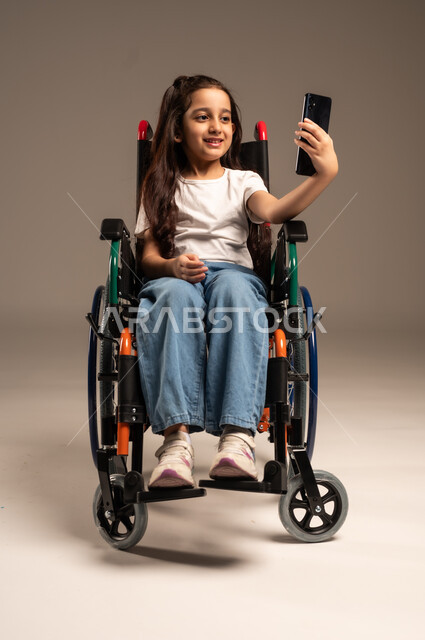 التقاط صورة سيلفي تذكارية، قضاء وقت ممتع بالتواصل مع الاصدقاء, التحدث مع الاقارب عبر الهاتف المحمول, بورتريه مقرب لفتاة عربية خليجية سعودية ترتدي الزي الكاجوال تجلس على كرسي متحرك تقوم بإجراء مكالمة فيديو, تأمين اجهزة ومعدات تساعد بالحركة والتنقل، خلفية رمادية