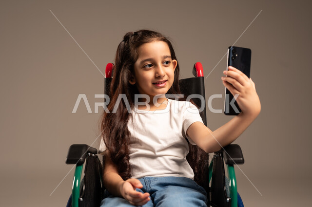 التقاط صورة سيلفي تذكارية، قضاء وقت ممتع بالتواصل مع الاصدقاء, التحدث مع الاقارب عبر الهاتف المحمول, بورتريه مقرب لفتاة عربية خليجية سعودية ترتدي الزي الكاجوال تجلس على كرسي متحرك تقوم بإجراء مكالمة فيديو, تأمين اجهزة ومعدات تساعد بالحركة والتنقل، خلفية رمادية