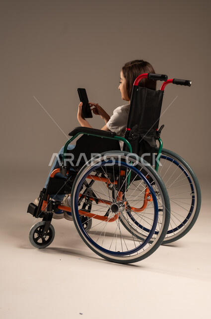 التقاط صورة سيلفي تذكارية، قضاء وقت ممتع بالتواصل مع الاصدقاء, التحدث مع الاقارب عبر الهاتف المحمول, بورتريه مقرب لفتاة عربية خليجية سعودية ترتدي الزي الكاجوال تجلس على كرسي متحرك تقوم بإجراء مكالمة فيديو, تأمين اجهزة ومعدات تساعد بالحركة والتنقل، خلفية رمادية