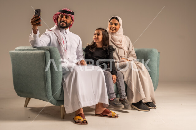 فرحة الأبناء بقضاء وقت ممتع مع الأهل, التقاط سيلفي عائلي تذكاري عبر الهاتف المحمول, بورتريه لعائلة عربية سعودية خليجية مبتسمة تقوم بتوثيق اللحظات الممتعة بإيماءات السعادة والفرح, الاجتماع الأسري في غرفة المعيشة وبناء مجتمع سعودي متنامي, خلفية رمادية