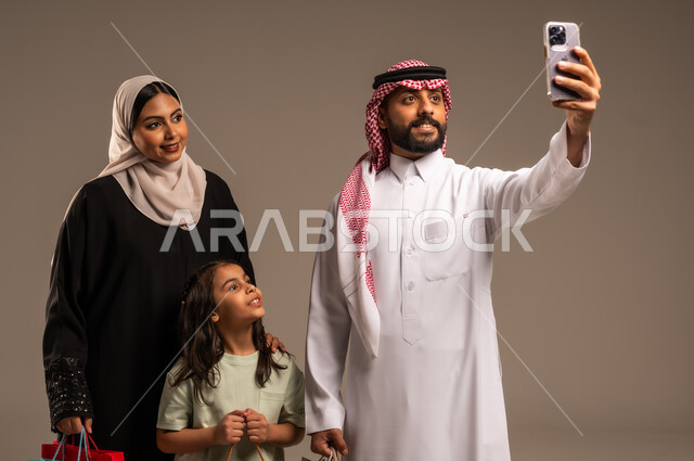 فرحة الأبناء بقضاء وقت ممتع مع الأهل, التقاط سيلفي عائلي تذكاري عبر الهاتف المحمول, بورتريه لعائلة عربية سعودية خليجية مبتسمة تقوم بتوثيق اللحظات الممتعة بإيماءات السعادة والفرح, الاجتماع الأسري في غرفة المعيشة وبناء مجتمع سعودي متنامي, خلفية رمادية