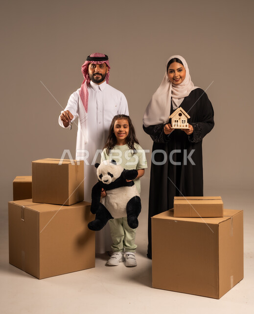 مفهوم الانتقال إلى سكن جديد، صناديق وحاويات كرتونية مليئة بالأغراض المنزلية، بورتريه لعائلة عربية سعودية خليجية تقف باستقامة بتعابير السرور، الاعتناء بالأطفال والاهتمام بهم, قضاء أوقات ممتعة مع الاسرة، الجسم بالطول الكامل، خلفيه رمادية
