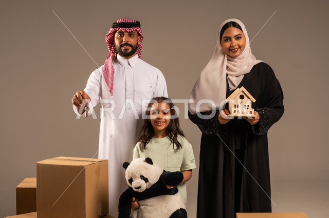 مفهوم الانتقال إلى سكن جديد، صناديق وحاويات كرتونية مليئة بالأغراض المنزلية، بورتريه لعائلة عربية سعودية خليجية تقف باستقامة بتعابير السرور، الاعتناء بالأطفال والاهتمام بهم, قضاء أوقات ممتعة مع الاسرة، الجسم بالطول الكامل، خلفيه رمادية