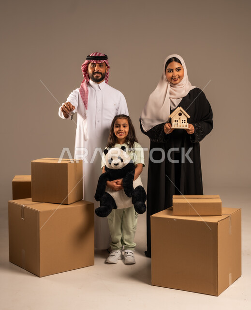 مفهوم الانتقال إلى سكن جديد، صناديق وحاويات كرتونية مليئة بالأغراض المنزلية، بورتريه لعائلة عربية سعودية خليجية تقف باستقامة بتعابير السرور، الاعتناء بالأطفال والاهتمام بهم, قضاء أوقات ممتعة مع الاسرة، الجسم بالطول الكامل، خلفيه رمادية
