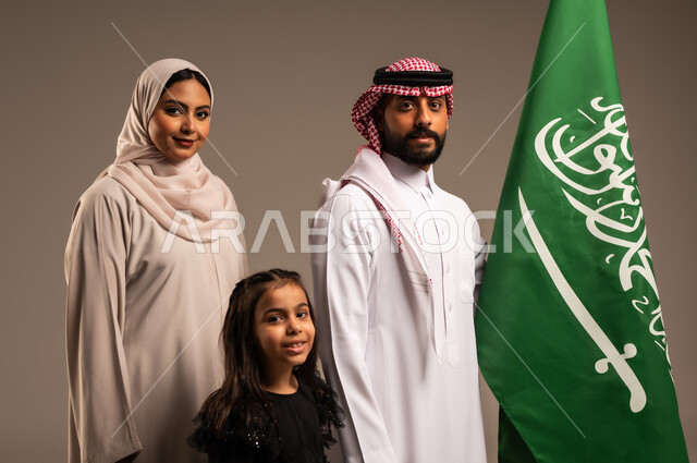 ذكرى اليوم الوطني السعودي، الأعياد والمناسبات الوطنية السعودية، الأجواء العائلية المليئة بالحب، بورتريه لعائلة عربية خليجية سعودية سعيدة تمسك بعلم السعودية بحب وفخر، الاحتفال بيوم العلم 11 مارس، العيد الوطني السعودي 8 ربيع الأول، خلفية رمادية