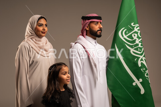 ذكرى اليوم الوطني السعودي، الأعياد والمناسبات الوطنية السعودية، الأجواء العائلية المليئة بالحب، بورتريه لعائلة عربية خليجية سعودية سعيدة تمسك بعلم السعودية بحب وفخر، الاحتفال بيوم العلم 11 مارس، العيد الوطني السعودي 8 ربيع الأول، خلفية رمادية