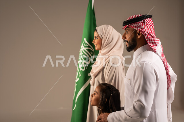 ذكرى اليوم الوطني السعودي، الأعياد والمناسبات الوطنية السعودية، الأجواء العائلية المليئة بالحب، بورتريه لعائلة عربية خليجية سعودية سعيدة تمسك بعلم السعودية بحب وفخر، الاحتفال بيوم العلم 11 مارس، العيد الوطني السعودي 8 ربيع الأول، خلفية رمادية