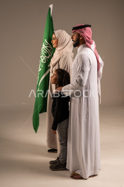ذكرى اليوم الوطني السعودي، الأعياد والمناسبات الوطنية السعودية، الأجواء العائلية المليئة بالحب، بورتريه لعائلة عربية خليجية سعودية سعيدة تمسك بعلم السعودية بحب وفخر، الاحتفال بيوم العلم 11 مارس، العيد الوطني السعودي 8 ربيع الأول، خلفية رمادية