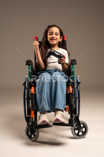 تحسين جودة حياة ذوي الاعاقة الحركية، تجربة مثيرة في الأجهزة الحديثة الالكترونية، نشاطات ترفيهية لذوي الهمم، التفاعل والاندماج مع ألعاب البلايستيشن, بورتريه لفتاة عربية خليجية سعودية ترتدي الزي الكاجوال تجلس على كرسي متحرك تحمل بيدها عصا التحكم،  خلفية رمادية