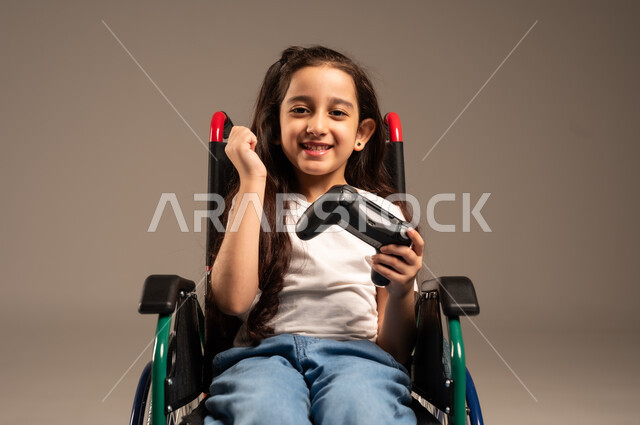 تحسين جودة حياة ذوي الاعاقة الحركية، تجربة مثيرة في الأجهزة الحديثة الالكترونية، نشاطات ترفيهية لذوي الهمم، التفاعل والاندماج مع ألعاب البلايستيشن, بورتريه لفتاة عربية خليجية سعودية ترتدي الزي الكاجوال تجلس على كرسي متحرك تحمل بيدها عصا التحكم،  خلفية رمادية