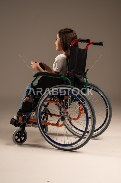 تحسين جودة حياة ذوي الاعاقة الحركية، تجربة مثيرة في الأجهزة الحديثة الالكترونية، نشاطات ترفيهية لذوي الهمم، التفاعل والاندماج مع ألعاب البلايستيشن, بورتريه لفتاة عربية خليجية سعودية ترتدي الزي الكاجوال تجلس على كرسي متحرك تحمل بيدها عصا التحكم،  خلفية رمادية